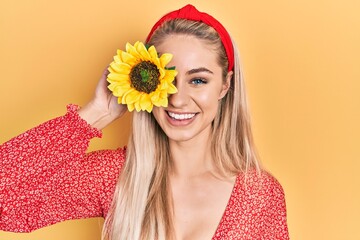 Poster - Young beautiful caucasian woman holding yellow sunflowers over eye looking positive and happy standing and smiling with a confident smile showing teeth