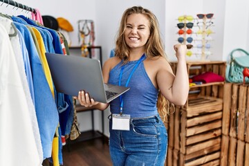 Wall Mural - Young caucasian woman working as manager at retail boutique holding laptop screaming proud, celebrating victory and success very excited with raised arms