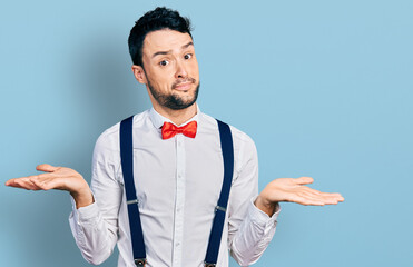 Poster - Hispanic man with beard wearing hipster look with bow tie and suspenders clueless and confused expression with arms and hands raised. doubt concept.