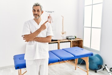 Sticker - Middle age hispanic therapist man working at pain recovery clinic pointing with hand finger to the side showing advertisement, serious and calm face