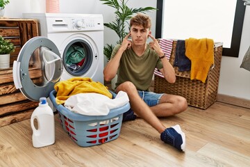 Canvas Print - Young caucasian man putting dirty laundry into washing machine covering ears with fingers with annoyed expression for the noise of loud music. deaf concept.