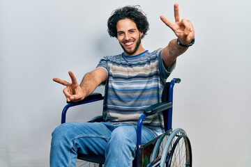 Canvas Print - Handsome hispanic man sitting on wheelchair smiling with tongue out showing fingers of both hands doing victory sign. number two.