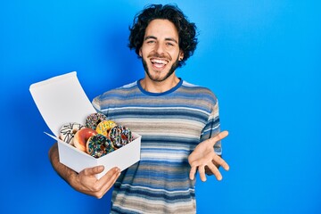 Wall Mural - Handsome hispanic man holding tasty colorful doughnuts celebrating achievement with happy smile and winner expression with raised hand