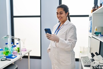 Poster - Young hispanic woman wearing scientist uniform using smartphone at laboratory