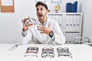 Sticker - Young optician man holding optometry glasses angry and mad screaming frustrated and furious, shouting with anger. rage and aggressive concept.