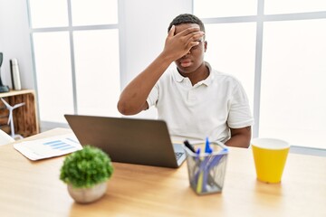 Wall Mural - Young african man working at the office using computer laptop covering eyes with hand, looking serious and sad. sightless, hiding and rejection concept