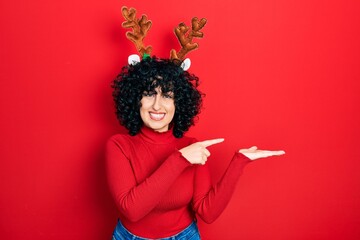 Poster - Young middle east woman wearing cute christmas reindeer horns amazed and smiling to the camera while presenting with hand and pointing with finger.