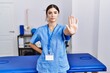 Young hispanic woman wearing physiotherapist uniform standing at clinic doing stop sing with palm of the hand. warning expression with negative and serious gesture on the face.