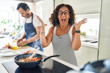 Middle age couple cooking mediterranean food at home celebrating victory with happy smile and winner expression with raised hands