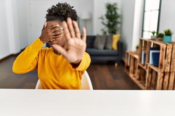 Poster - Young african american woman wearing casual clothes sitting on the table at home covering eyes with hands and doing stop gesture with sad and fear expression. embarrassed and negative concept.