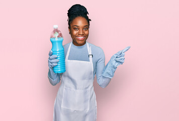 Sticker - Young african american woman wearing apron holding detergent bottle smiling happy pointing with hand and finger to the side