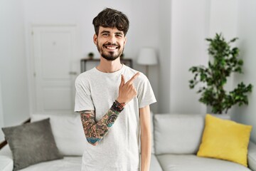 Wall Mural - Hispanic man with beard at the living room at home cheerful with a smile on face pointing with hand and finger up to the side with happy and natural expression