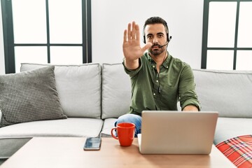 Wall Mural - Young hispanic man with beard wearing call center agent headset working from home doing stop sing with palm of the hand. warning expression with negative and serious gesture on the face.