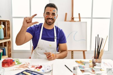 Poster - Young hispanic man with beard at art studio with painted face smiling and confident gesturing with hand doing small size sign with fingers looking and the camera. measure concept.