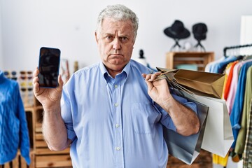 Sticker - Senior man with grey hair holding shopping bags and showing smartphone screen relaxed with serious expression on face. simple and natural looking at the camera.