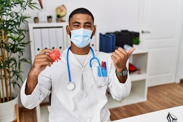 Poster - Young indian doctor holding virus toy wearing safety mask pointing thumb up to the side smiling happy with open mouth