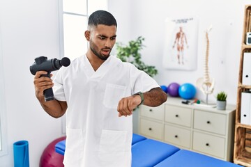 Wall Mural - Young indian physiotherapist holding therapy massage gun at wellness center looking at the watch time worried, afraid of getting late