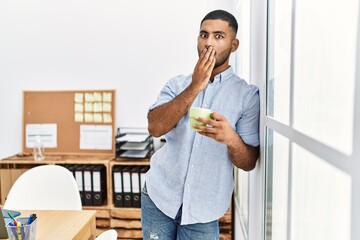 Poster - Young indian man drinking a cup coffee at the office covering mouth with hand, shocked and afraid for mistake. surprised expression