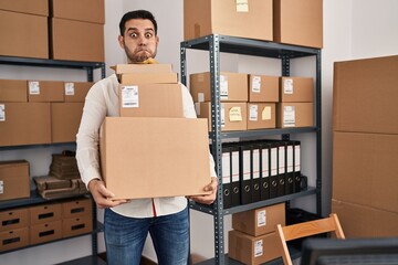 Sticker - Young hispanic man with beard working at small business ecommerce holding delivery boxes puffing cheeks with funny face. mouth inflated with air, catching air.