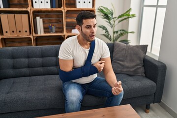 Canvas Print - Young hispanic man with beard wearing arm on sling sitting at therapy consult scared and amazed with open mouth for surprise, disbelief face