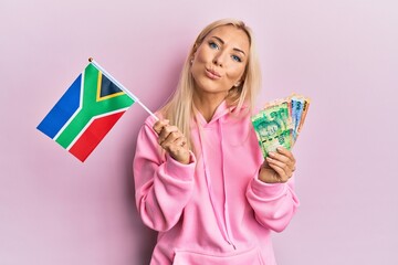 Wall Mural - Young blonde woman holding south africa flag and rand banknotes looking at the camera blowing a kiss being lovely and sexy. love expression.