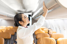 Asian Female Flight Attendant Closing The Overhead Luggage Compartment Lid For Carry On Baggage After Passengers Are Seated And Prepare To Take Off