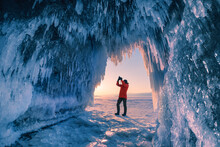 Man Tourist Photograph The Ice Cave On Baikal Lake At Sunset. Winter Landscape Of Baikal Lake, Siberia, Russia