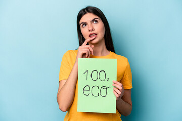 Young caucasian woman holding 100% eco placard isolated on blue background relaxed thinking about something looking at a copy space.