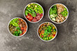 4 poke bowls with tofu, salmon, chicken, tuna, cucumbers, salad leafs, edamame beans, spinach and peanuts on a gray textured background. Top view.