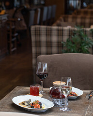 On the table in the restaurant serving for 2 persons. Dish with fish and eggplant, a glass teapot with red tea, a mug of tea, a glass of white and red wine, a plate with carpaccio in the background.