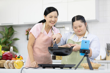 mother and down syndrome teenage girl or her daughter cooking food together and live streaming online via smartphone on tripod in a kitchen