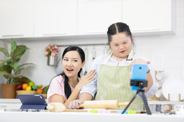 mother and down syndrome teenage girl or her daughter rolling out dough and live streaming online via smartphone on tripod in a kitchen