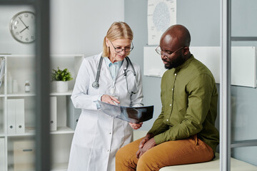 Poster - Confident mature doctor in whitecoat showing young black man x-ray image of his lungs and explaining him diagnosis in clinics