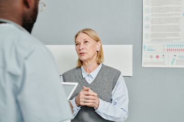 Poster - Contemporary mature female patient in casualwear describing her symptoms to doctor while sitting in front of him in hospital