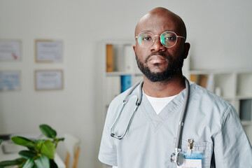 Wall Mural - Young contemporary African American male clinician in eyeglasses and blue uniform standing in medical office and looking at camera