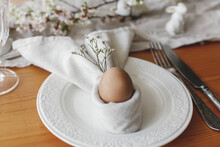 Stylish Easter Brunch Table Setting. Easter Egg In Bunny Napkin On Plate With Cutlery, Bunny, Spring Flowers And Rustic Cloth On Wooden Table. Easter Table Arrangement And Eco Friendly Decorations