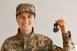 Portrait of smiling positive optimistic military woman wearing camouflage uniform and cap, standing looking at camera with happy smile and holding keys from a new car or apartment.