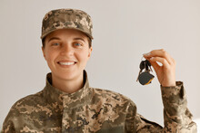 Portrait Of Smiling Positive Optimistic Military Woman Wearing Camouflage Uniform And Cap, Standing Looking At Camera With Happy Smile And Holding Keys From A New Car Or Apartment.