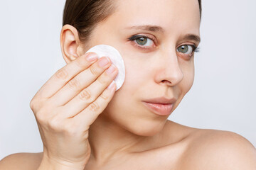 Wall Mural - Close up of young caucasian woman cleaning her face with lotion using cotton pad isolated on a white background. Skin care, cosmetology. Evening routine. The girl washes off her makeup with a cleanser