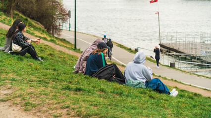 Girls rest on the green grass on the banks of the river on a cloudy day in early spring. Back view without a face