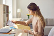 © Studio Romantic - Young woman opens paper letter that she received by mail. Side view of student girl sitting at working desk with laptop computer at home and taking notification document out of envelope