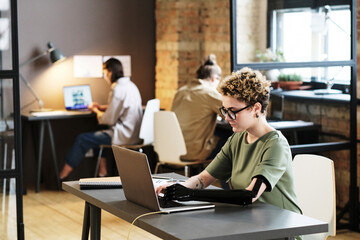 Young woman in eyeglasses with prosthetic arm typing on laptop during her work at office