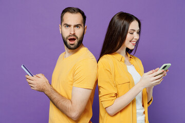 Wall Mural - Side view young indignant jelaous couple two friends family man woman together in yellow clothes hold in hand use mobile cell phone stand back to back look camera isolated on plain violet background.