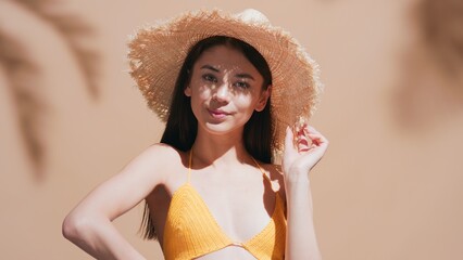 Wall Mural - Close-up beauty portrait of young brown-haired woman in a straw hat enjoys the sun against a beige background | Sunscreen cream promotional concept