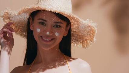 Wall Mural - Young brown-haired asian woman in a straw hat applies some sunscreen on her nose, enjoys the sun and smiles for the camera against beige background | Sunscreen commercial