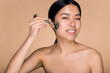 © Kateryna - Charming beautiful young Asian woman with naked shoulders, using derma roller for massage her facial skin. Photo of a smiling chinese girl on isolated beige background, cares for the skin