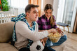 © Miljan Živković - two people couple man and woman boyfriend and girlfriend or husband and wife sitting on the sofa at home watching football soccer game cheering copy space eat popcorn and hold the ball