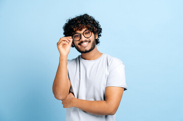 Attractive handsome positive indian or arabic trendy curly-haired guy with glasses, freelancer or creative designer, stands on a blue isolated background, looks at the camera, smiles friendly