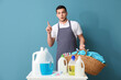 Young man with laundry basket, clothespins, table and bottles of detergent on blue background