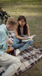 © iaginzburg - Sitting on the plaid in a public park, a young asian girl with long dark hair is reading a book for her disabled friend in a blue shirt